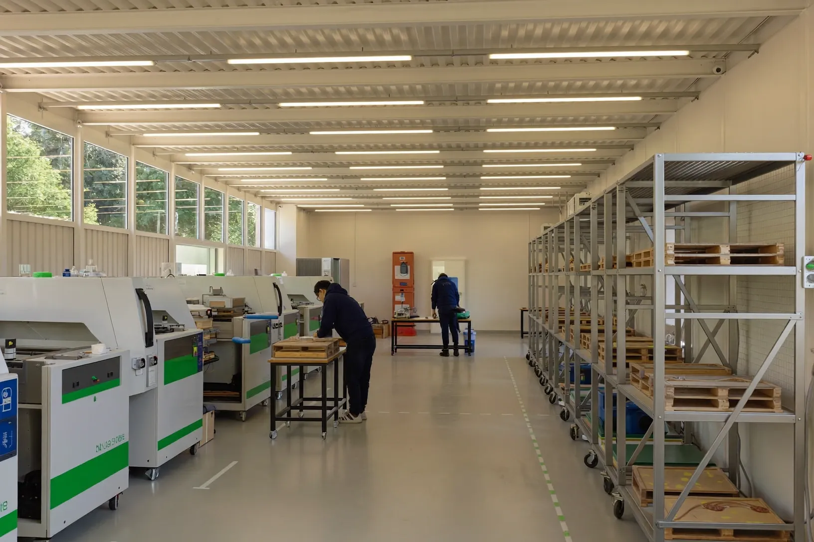 Eine von Neonlicht beleuchtete Produktionshalle. Zwei Personen arbeiten entlang einer Reihe von Maschinen gegenüber Regalen mit gestapelten Holzrahmen.